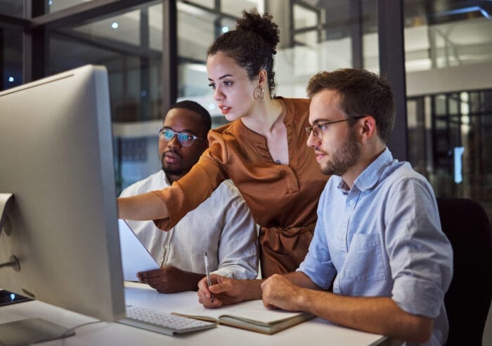 Three professionals collaborating on a project in an office setting, with one pointing at a computer screen and the others attentively engaged